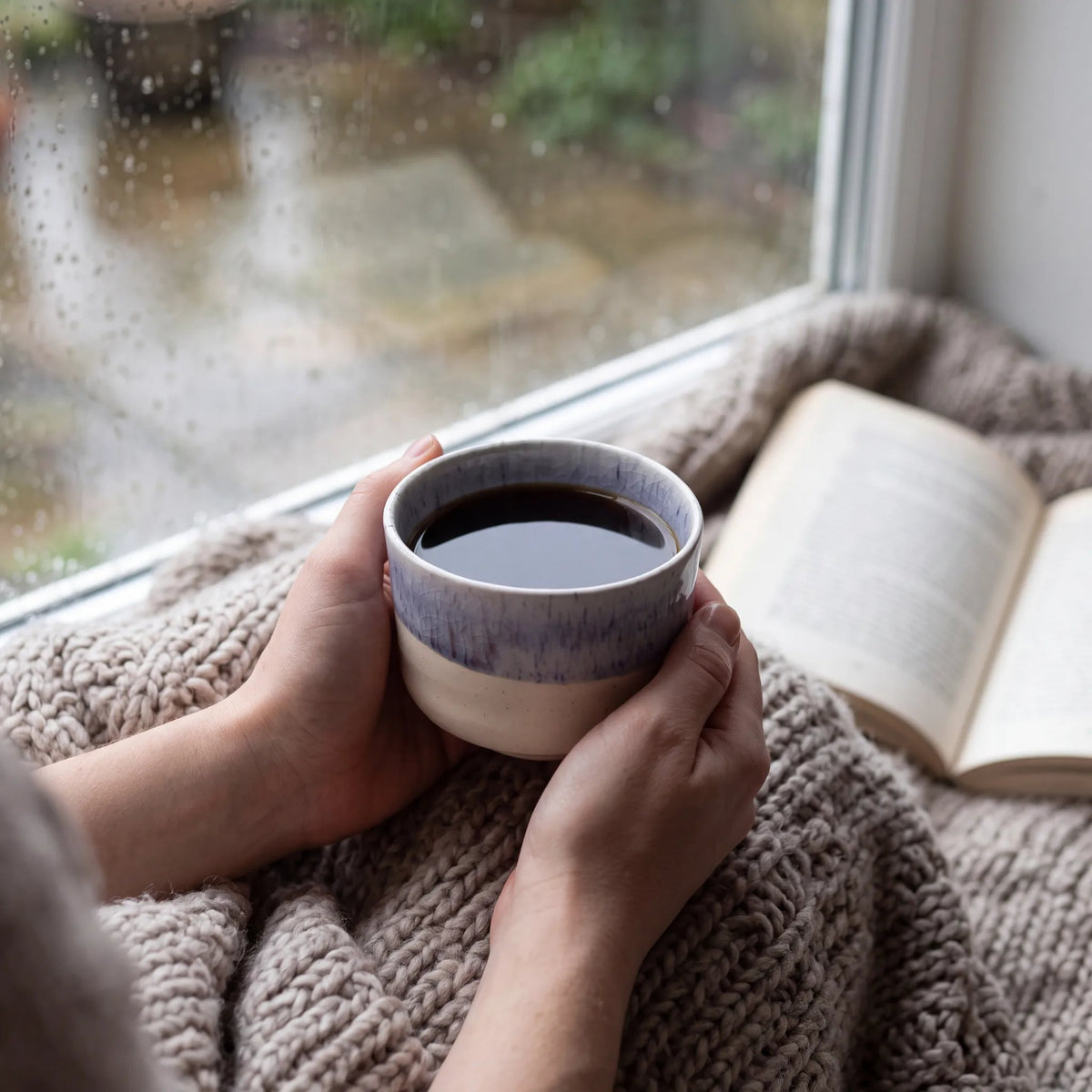 Winter Drift koffiekop in twee handen op een deken, met een boek bij het raam